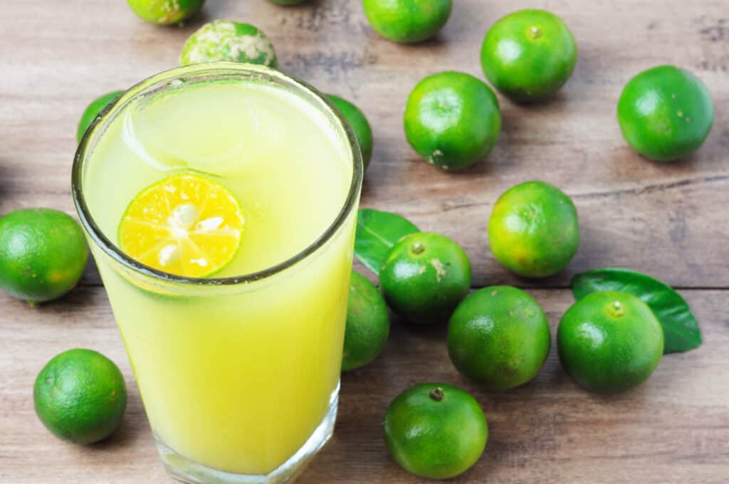 A tall clear glass filled with yellow-green Calamansi juice with a thin, circular slice of calamansi floating in it. The glass is on a wooden table that also contains a dozen or so whole bright green calamansi fruits.