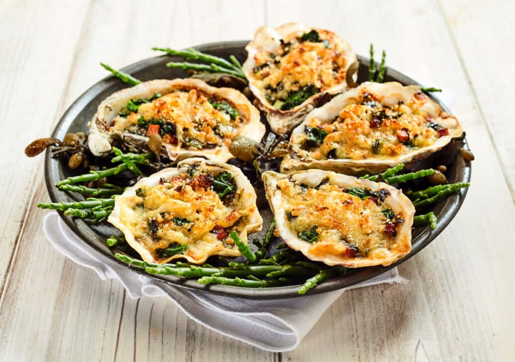 five oysters served on the half shell on. a round plate, center frame. The oysters have been cooked and are covered with melted Parmesan cheese. Green garnish adorns the plate.