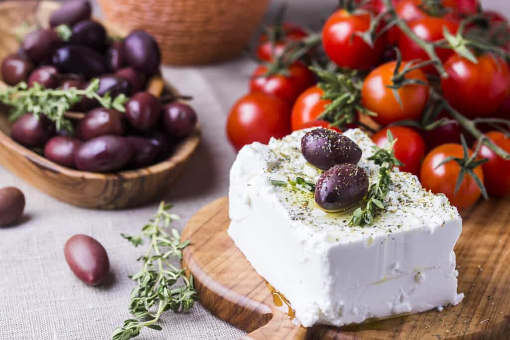 a rectangle of greek feta cheese on a wooden cutting board. The cutting board is on a light-colored cloth covered table. The feta has two ripe purple/black olives on top of it along with a sprinkle of finely ground black pepper a sprig of thyme. Another sprig of time is loose on the table, along with two ripe olives. In the background (frame left) is a shallow wooden bowl heaped with ripe olives. Behind the cheese (upper right frame) are a dozen or so small (cherry) orange tomatoes, still attached to the vine, which is drying. 