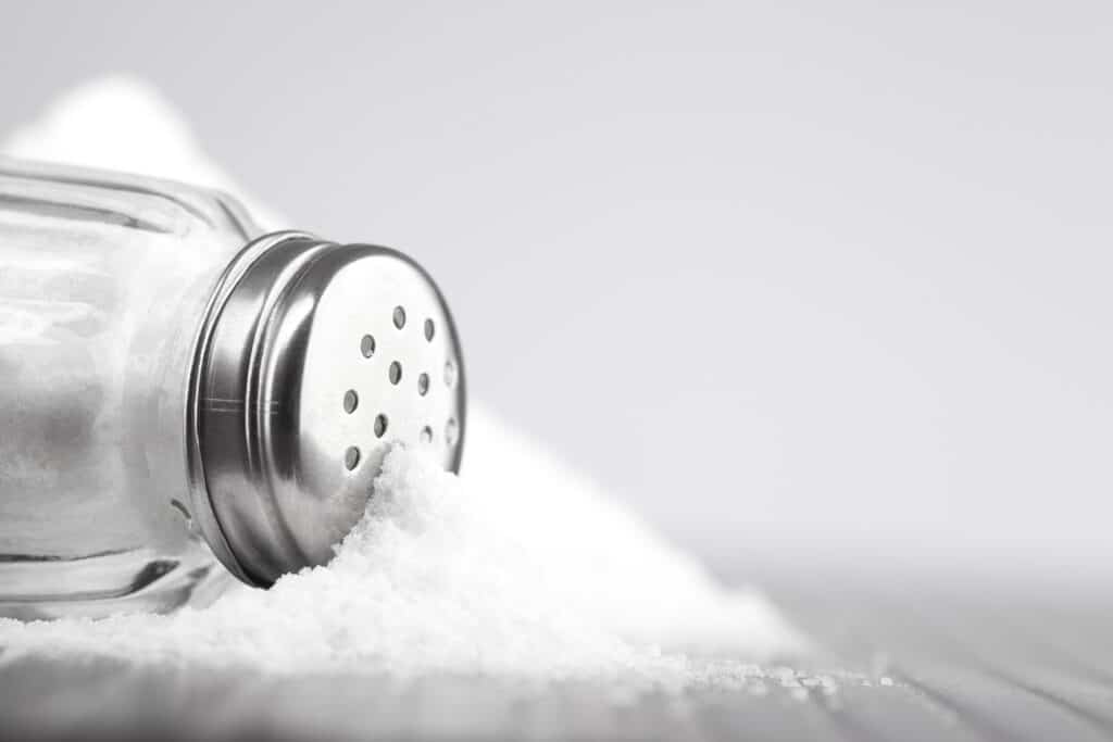 Frame Left: horizontal salt shaker on its side, in a mound of salt that is low center frame. The salt shaker is glass with a stainless steel top with a a dozen visible holes.