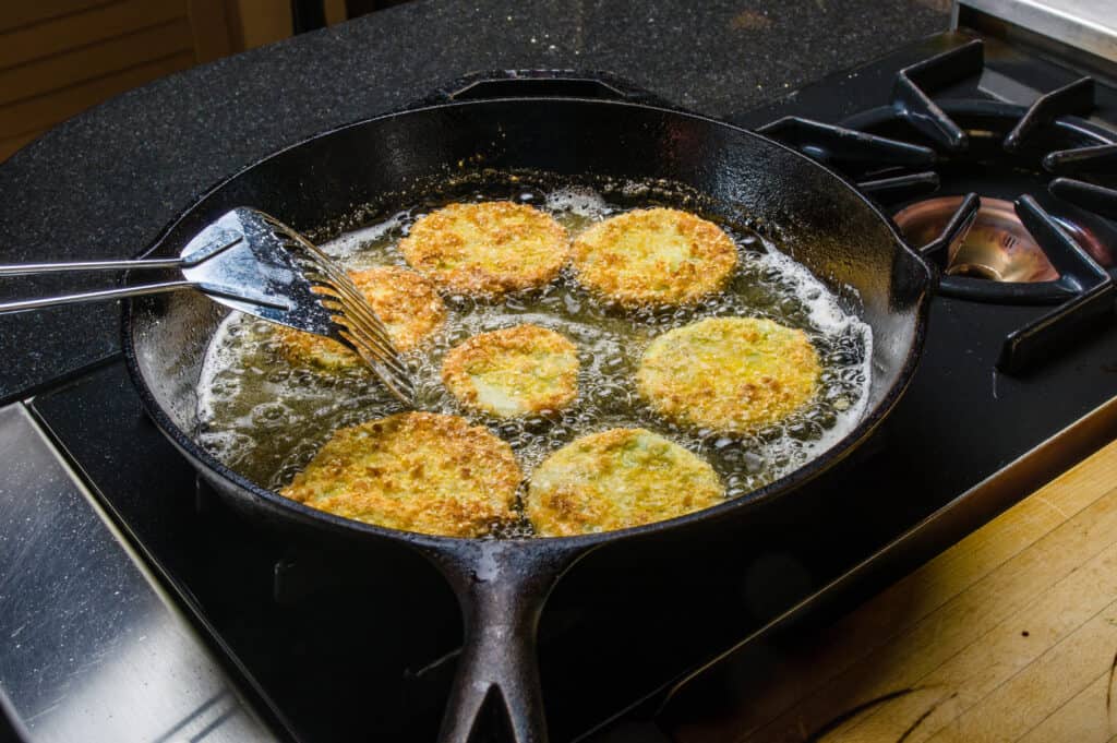 High slices of green tomato coated in corn meal, frying  in oil in a cast iron skillet odn a gas stove. 