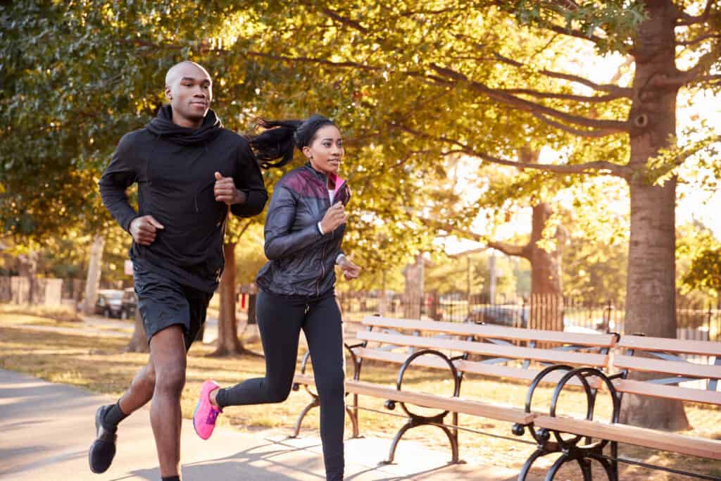 Two humans jogging on pavement passed a series of natural wooden benches with black metal armrests in park-like setting with many leafy trees and grass.