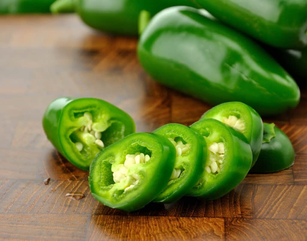 sliced up green jalapeno peppers with white seed on a brown wooden table. three green jalpenos are on the table behind the slices, of which there are four, with the ends of the pepper they were sliced from visible nearby.