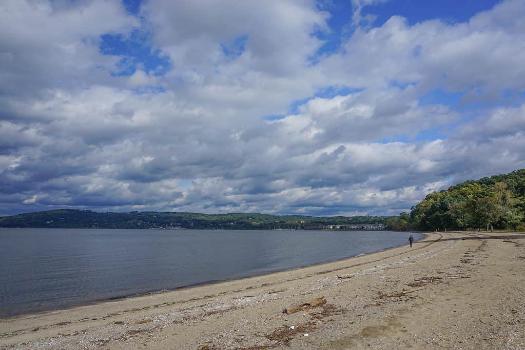 Croton-On-Hudson, New York, USA: A solitary person walks along the Hudson River under a cloud-filled sky, on a pebble beach in Croton Point Park.