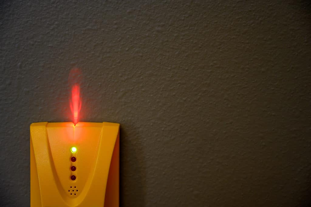 The background is a charcoal-black wall isolate: bottom frame left is a plastic bright orange electronic stud finder. There are four round lights in the center of the stud finder the top one is illuminated green. Below the lights is a circle composed of six small holes with one small hole in the center. A bright red flame like light is shooting out of the top of the stud finder onto the charcoal-black wall.