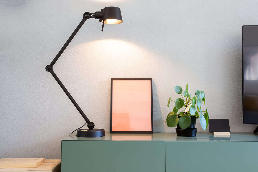 A Chinese money plant in a round black pot with a matching saucer is visible center-frame right.. The plant is smallish . with about 15 round green leaves. It is sitting on a green desk or table with a black desk lamp with a hinged arm attached to a round base is visible to the left . an empty picture frames leaning agains the gray wall. The light is shining on the frame and the plant. The edge of a flat screen television or monitor is barely visible, frame right.