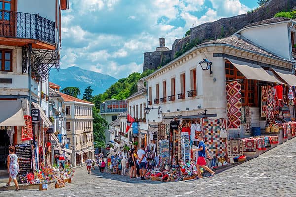 Gjirokaster town, Albania –August 2, 2020: summer cityscape – street of ancient town with souvenir shops and tourists. Old historical architecture and Castle complex on the hill.