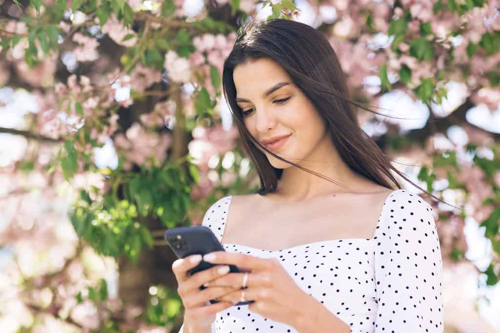 桜をバックにスマートフォンを操作している女性の写真