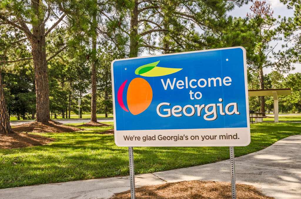 A welcome to Georgia sign in front of a rolling meadow with trees in the background.