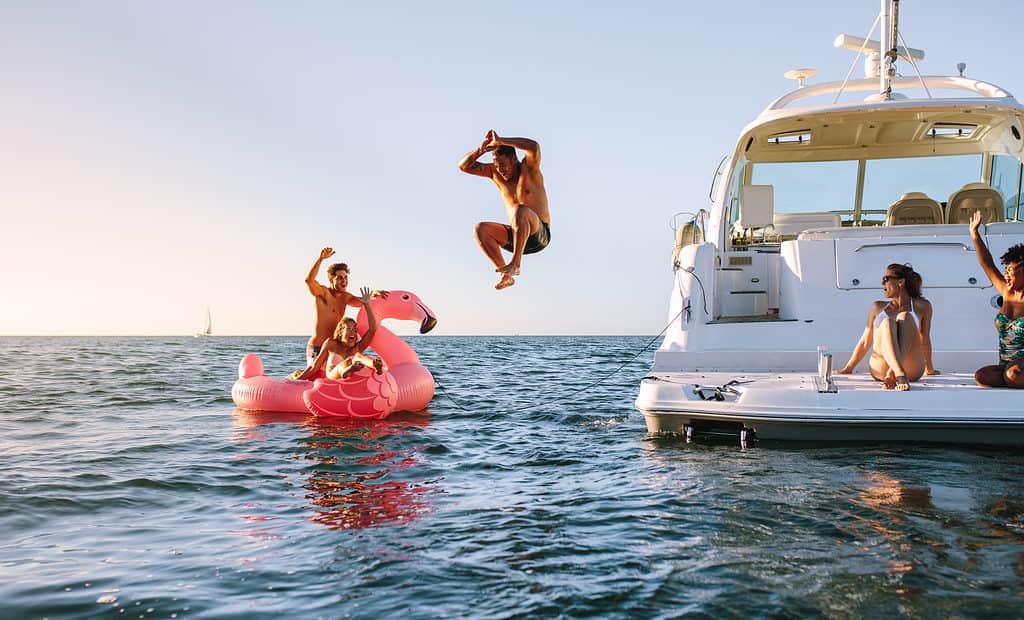 Photo of people having a lot of fun on a lake. There is a sailboat in the distance at the left at the horizon line. In the left frame there are two people on a rather large pink flamingo float. They seem to be cheering on a person who has jumped off the boat is and is in mid air. The person in mid air is in the center of the frame. The right frame consists of a modern-looking white pleasure boat on which two people are sitting and watching the person who has jumped from the boat. All of the people are tanned light-skinned people.