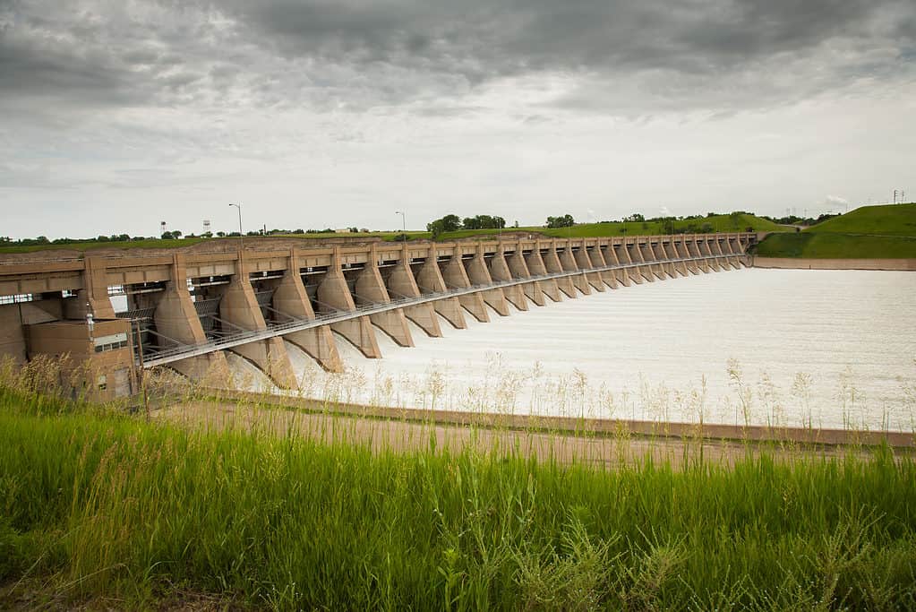 Photo of a dam on a river. The dam is long and made of concrete. IThere is water pouring through the dam. in the foreground green tall prairie grass is growing 