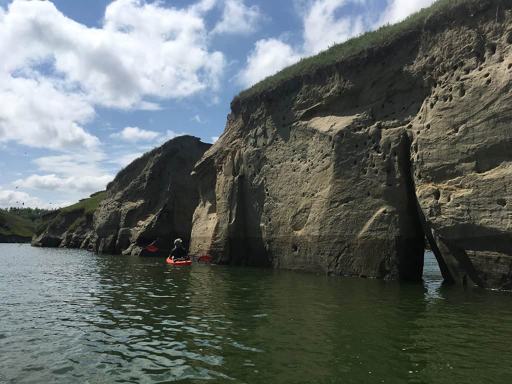 An orange boat is seen in the center frame but a little far off in a lake that has an enormous rock Cliff on the right the sky blue with white clouds as seen in the background as well as an embankment with some Prairie grass growing on it. The water takes up the foreground and is a deep green with a slightly choppy surface as if a light wind or distant boat is at play.