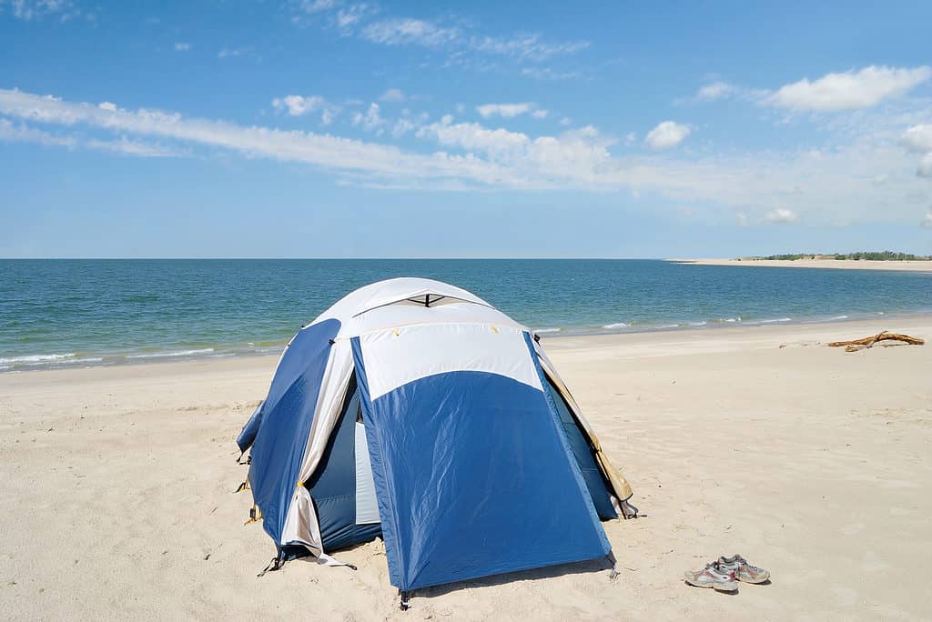 Photo of a blue and silver dome-type tent on a white sand lake beach. There is a pair of lace-up running shoes visible near the tent. The lake and a blue sky with a few wispy white clouds make up the background.