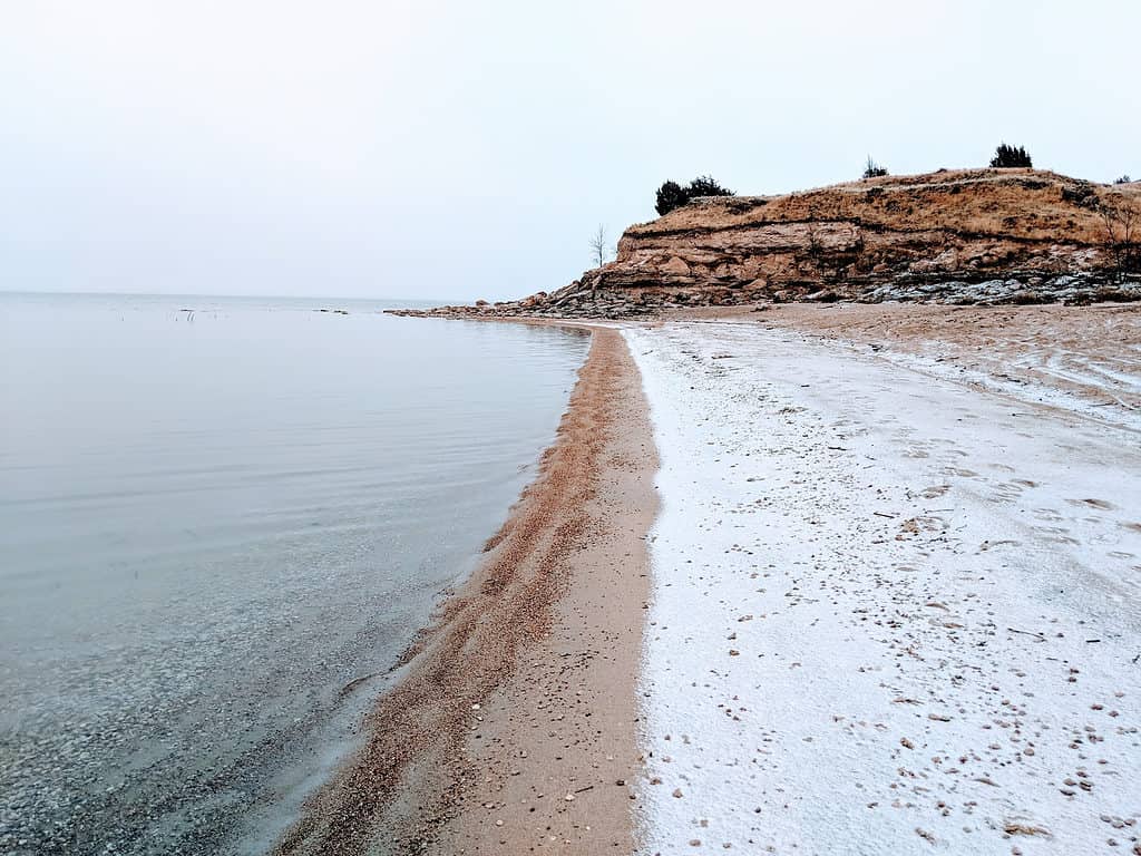 A photograph of a sandy shoreline, with a thin coat of snow. There is an outcropping  in the background. The lake is in the left frame, the beach/shoreline takes up frame right