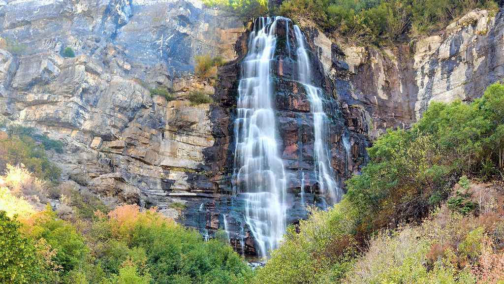 Photograph of Bridalveil falls in Provo canyon Utah. The falls are slightly right of center frame and appear to be white on a dark brown to light brown rock. There is a rock ledge on either side of the falls that is lighter brown because it is not wet. In the foreground of the photograph is shrubby green to brown vegetation above the Falls is the same. No sky is visible.