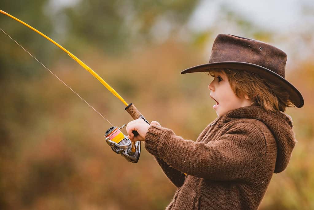 A young child dressed in a long sleeve brown knitted sweater. The child is a wearing a leather wide-brim hat. The child is holding a yellow rod and reel (fishing pole). The child is facing frame left and the expression on their face appears to be one of elation as if they have just caught a large fish. The background is out of focus nature