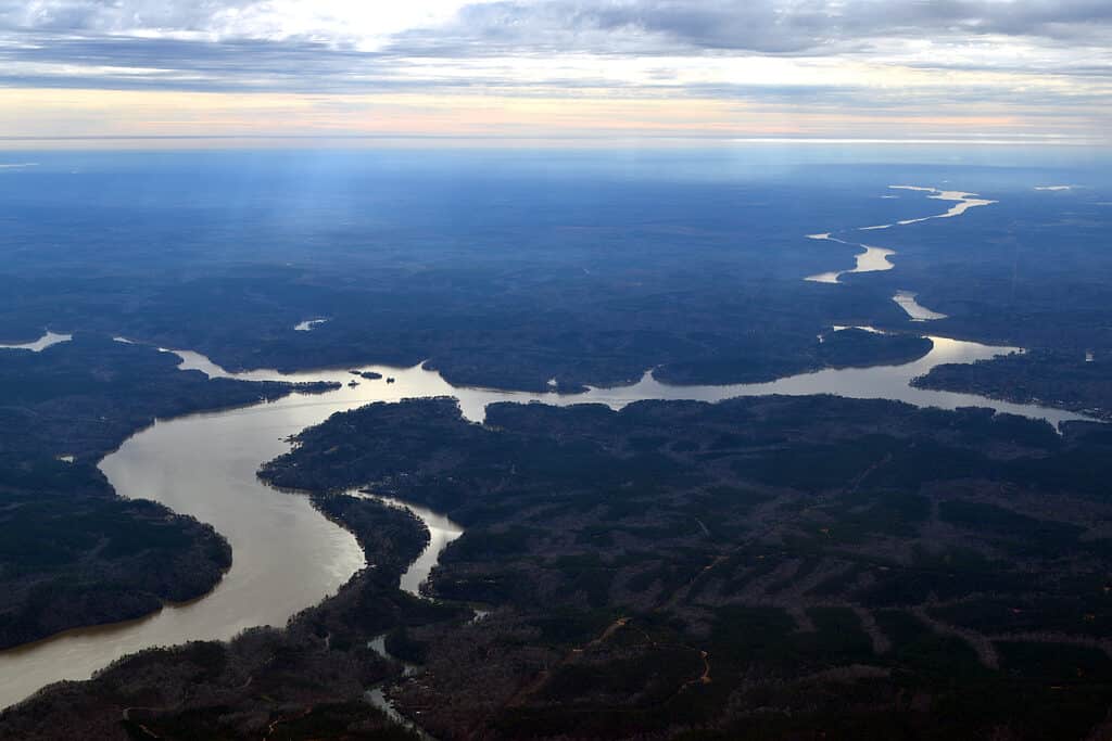 An aerial view of the Coosa River in Alabama. The river is visible meandering through the Fram at a diagonal fro the top right frame to the bottom left frame. The river is surrounded by indistinct vegetation. 