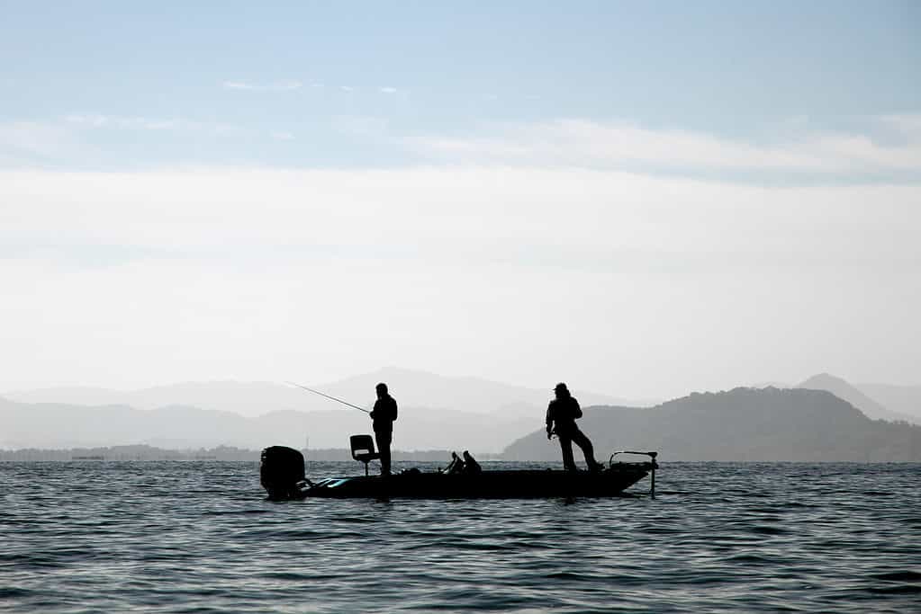 Center frame of photo: Dark silhouette of two men standing up fishing in a small outboard motor boat on a lake.