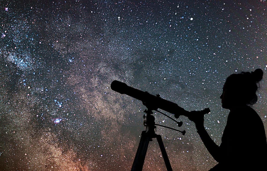 Photo lower right frame silhouette of a person with their hair in a top knot. The person’s right hand is clutching a telescope also silhouette, against a star filled sky