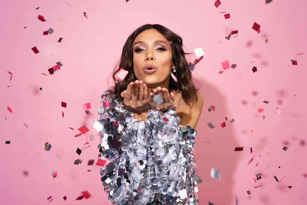 Woman in shiny silver outfit blowing away confetti, pink background