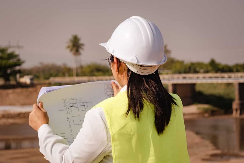 Engineer wearing a helmet and safety vest works and looks at the blueprints for construction plan and design details of the dam.