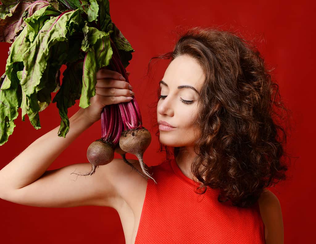Pretty light-skinned brunette young sport woman posing with fresh beetroot green leaves. Dieting. Healthy eating concept on red background