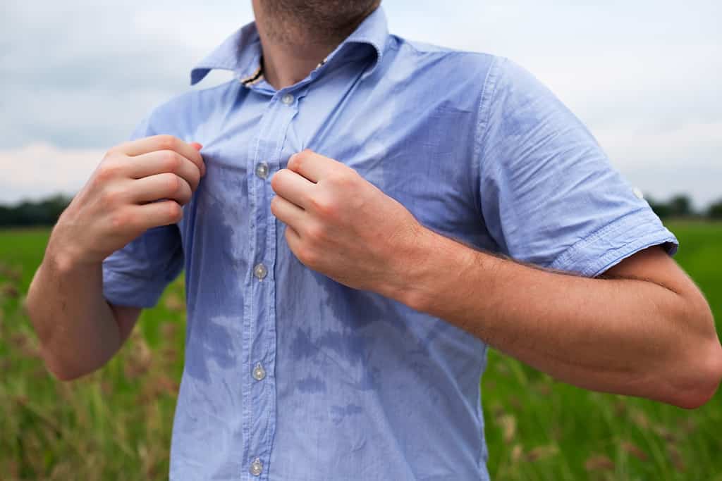 Man with hyperhidrosis sweating very badly under armpit in blue shirt because of hot weather. Travelling in asia thailand with backpacker. The man is pulling at his shirt, as if to fan himself with it. The shirt is visibly wet with sweat. The man is tanned. He is standing in a field. All that is visible is his chin and torso. 