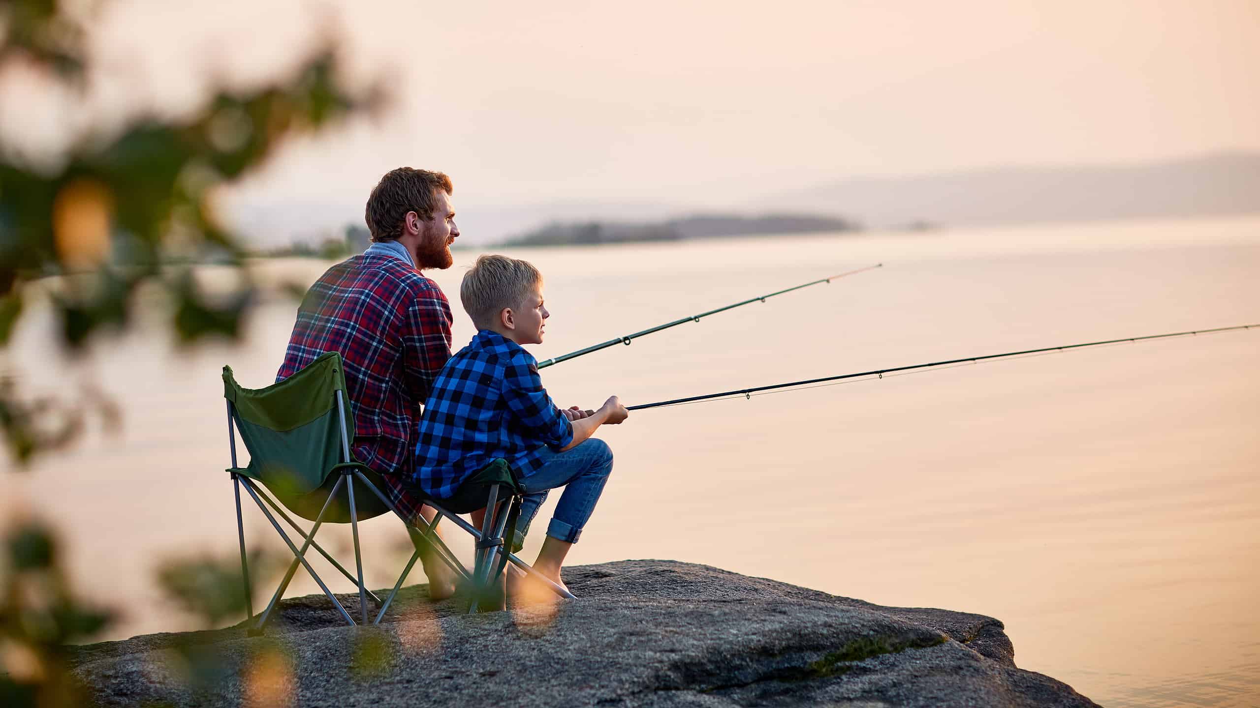Side view portrait of father and son sitting together on rocks fishing with rods in calm lake waters with landscape of setting sun, both wearing checkered shirts, shot from behind tree