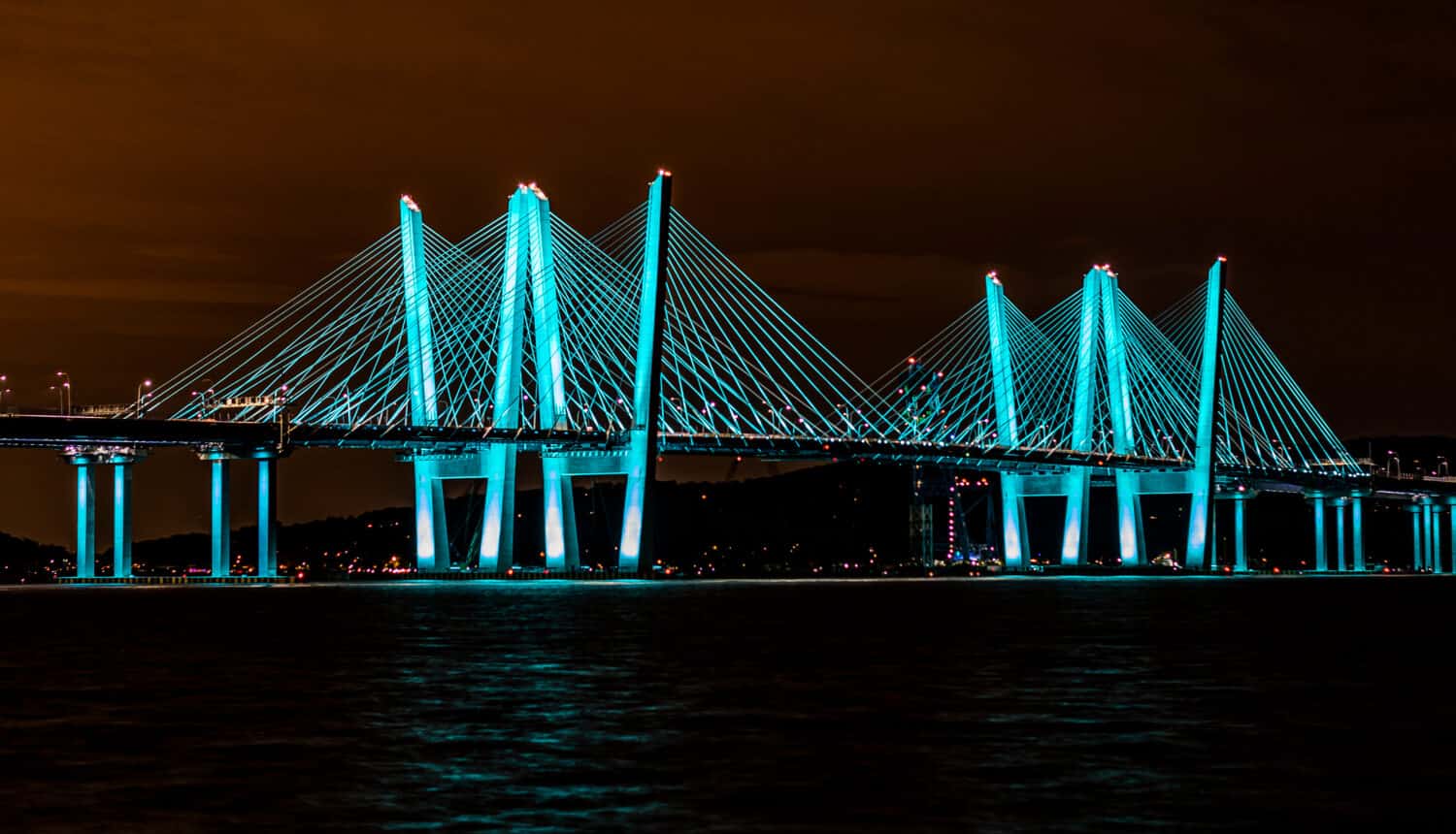 View of the Governor Mario M. Cuomo Bridge (new Tappan Zee Bridge) at night from Pierson Park in Tarrytown, NY.   The bridge is colored in a beautiful blue green hue with a fiery night sky.