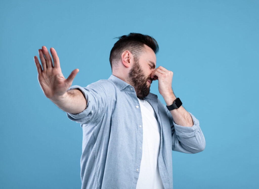 Bad smell concept. Young bearded guy closing his nose and showing STOP gesture, expressing disgust on blue studio background. Millennial man repulsed by toxic odor or terrible perfume