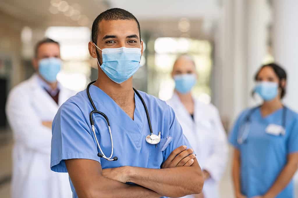 Confident multiethnic male nurse in front of his medical team looking at camera wearing face mask during covid-19 outbreak. Happy and proud indian young surgeon standing in front of his colleagues.