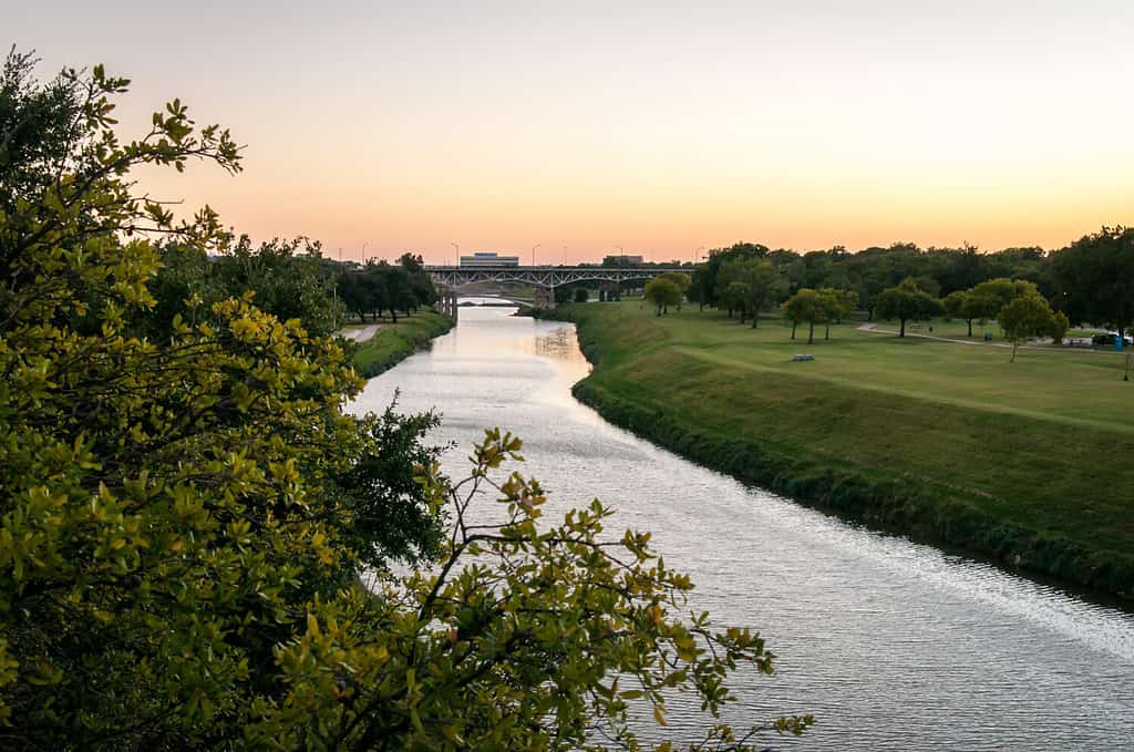 Trinity River Fort Worth, Texas. Sunset over the water, orange and golden colors. Green grass and trees in a public park. Peaceful and calm summer evening. Horizontal orientation.