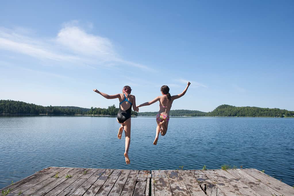 The backs of two young people holding hand are visible jumping off a wooden dock into a lake. 