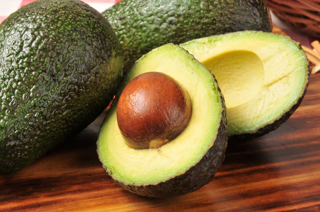 An avocado that has been sliced in half s visible on a wooden cutting board. The pit is still in the half that is closest to the lens. Two whole, uncut avocados are visible in the frame behind the sliced avocado.