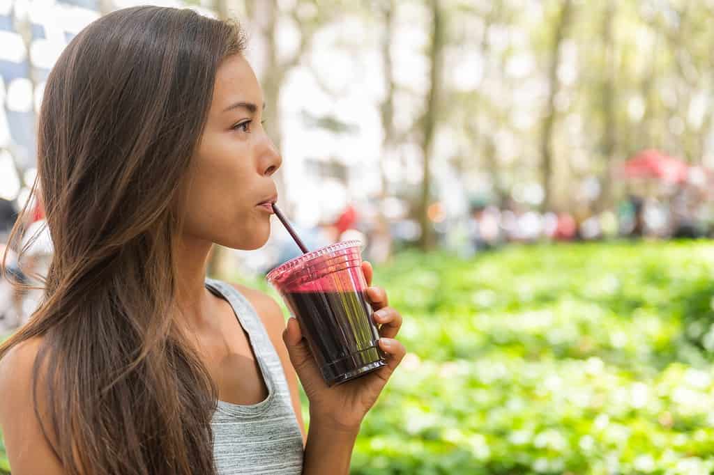 Olive-skinned woman with high cheekbones and long dark hair, wearing a sleeveless grey t-shirt with muted stripes, is visible in the left frame drinking beet juice smoothie walking in city park.