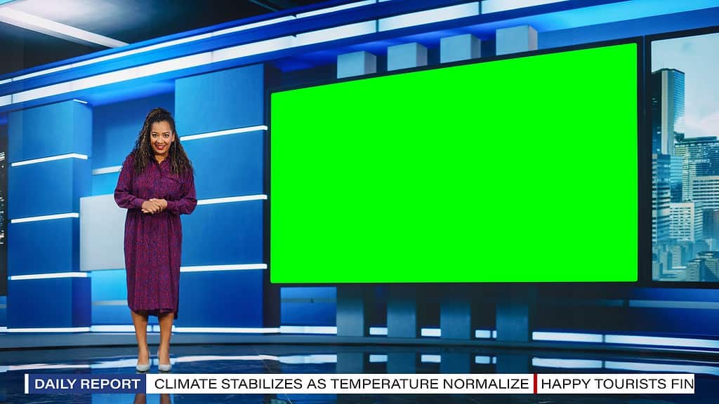 Brown-skinned Female Presenter Standing in Newsroom Studio, Uses Big Green Chroma Key Screen. News Achor, Host Talks about Weather.  The woman is in the left frame. A blank "green screen" is to her right.