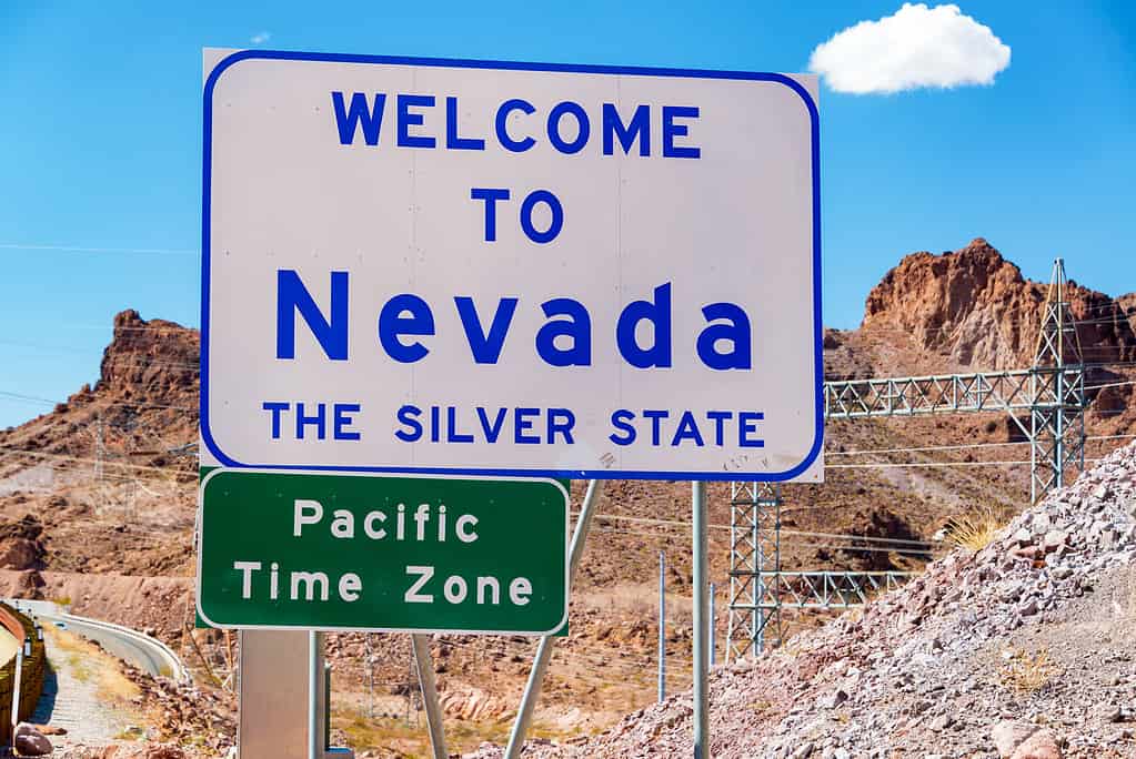 Welcome to Nevada sign on the border of Nevada and Arizona near Hoover Dam, The sign is white with blue lettering. A small green sign in the lower left frame says "Pacific Time Zone" in white letters. 