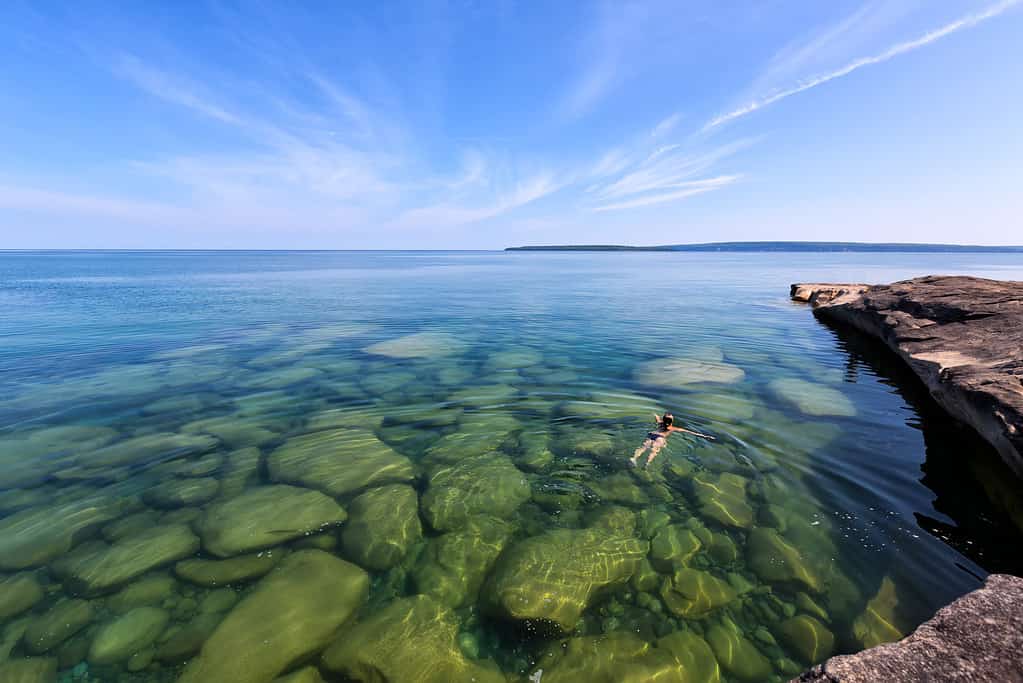 A girl swims in Lake Superior in the upper peninsula of Lake Michigan. Rocks are visable through the glass like, pristine waters. Pictured Rocks National Lakeshore is in the distance.