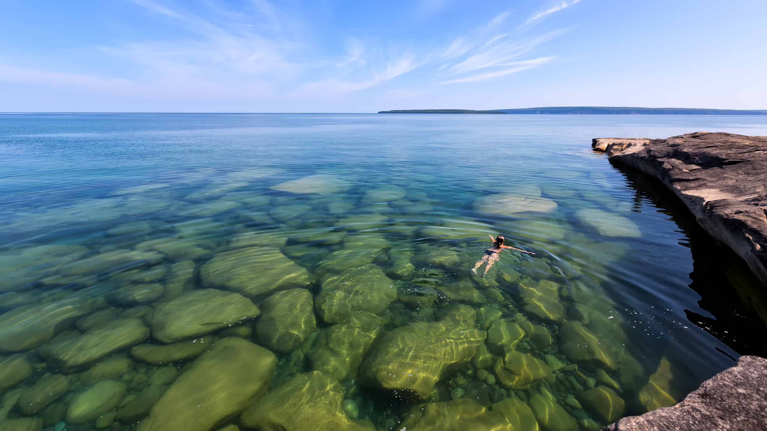 A girl swims in Lake Superior in the upper peninsula of Lake Michigan. Rocks are visable through the glass like, pristine waters. Pictured Rocks National Lakeshore is in the distance.