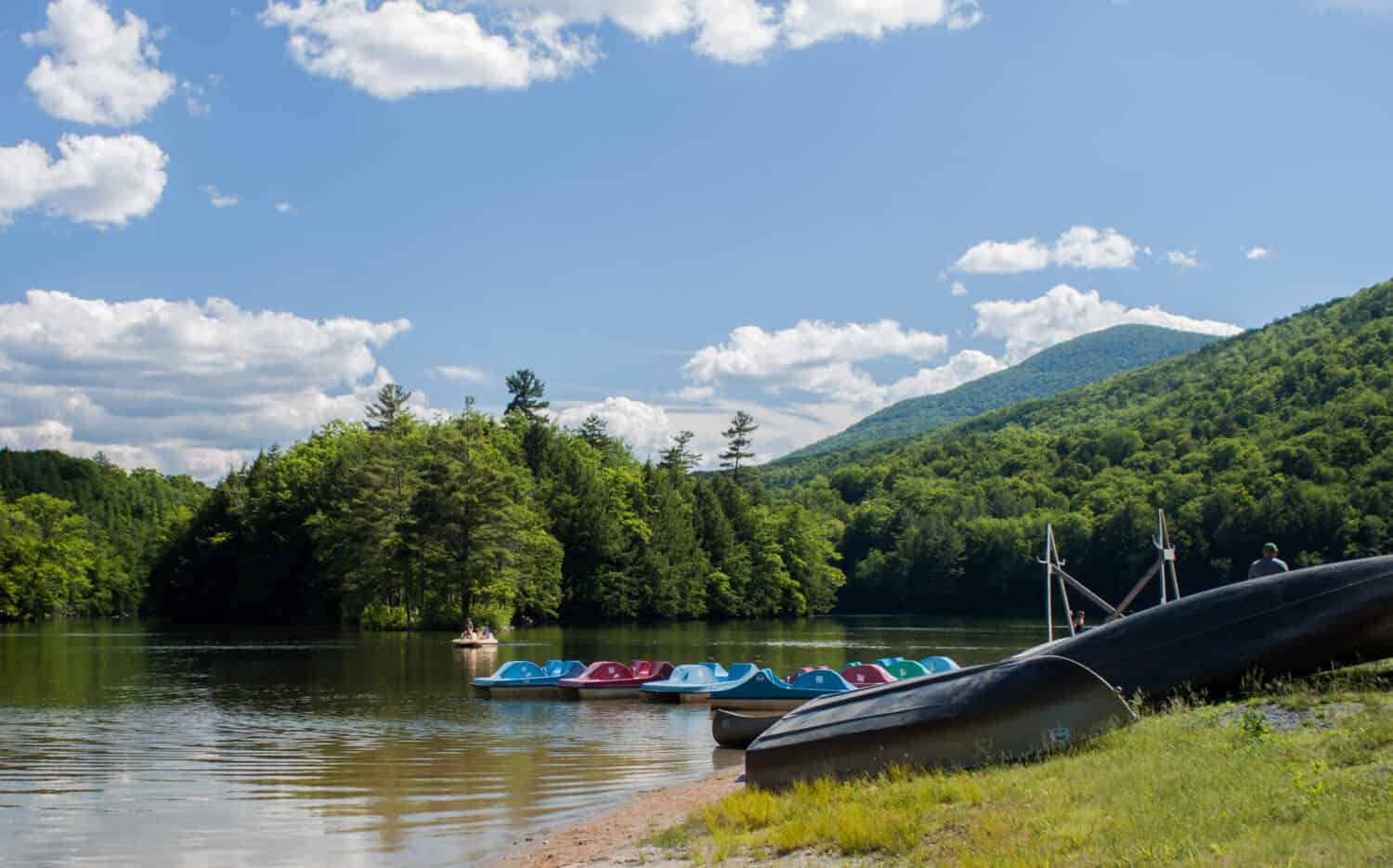 A row of paddle boats and canoes near the shore on Emerald Lake at Emerald Lake State Park in East Dorset, Vermont. The lake is surrounded by green trees and the sky is clear blue with a few clouds.