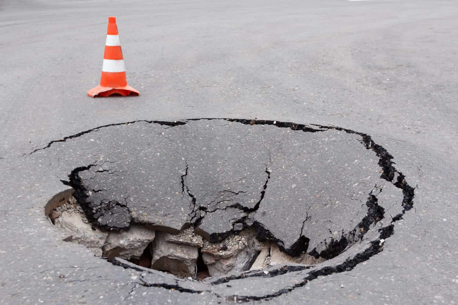 Deep sinkhole on a street city and orange traffic cone. Dangerous hole in the asphalt highway. Road with cracks. Bad construction. Damaged asphalt road collapse and fallen. 