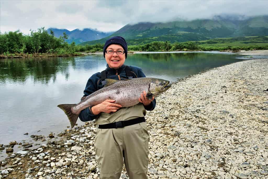 A happy, smiling fisherwoman holds a large King salmon that she caught after a long fight. She is standing on a rocky bank of a remote river in the mountains of Alaska.
