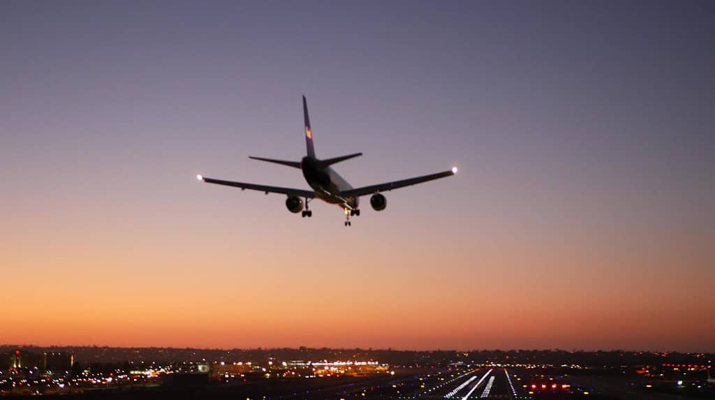 Airport runway lights at night, plane or airplane landing to airstrip, twilight dusk and sunset. Airliner jet arriving to aerodrome, San Diego airfield, California USA. Aircraft flying mid air in sky.