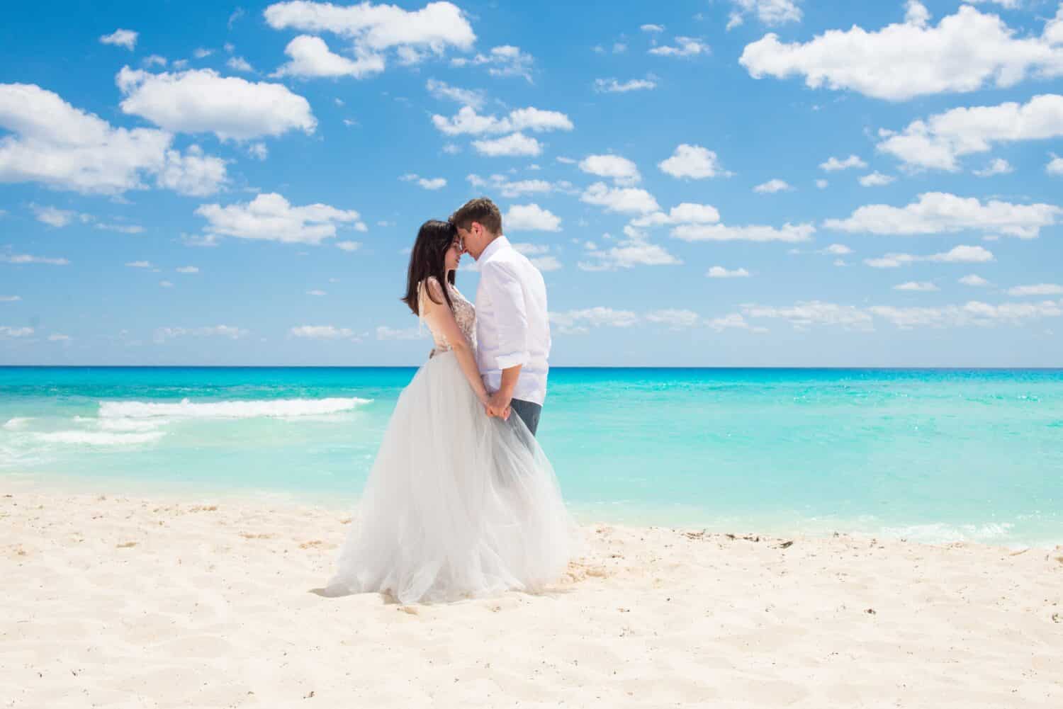 A married couple holding their hands and looking into their eyes. A wedding on the beach-shore. Honey moon on the paradise island. White sand, turquoise sea waters. 