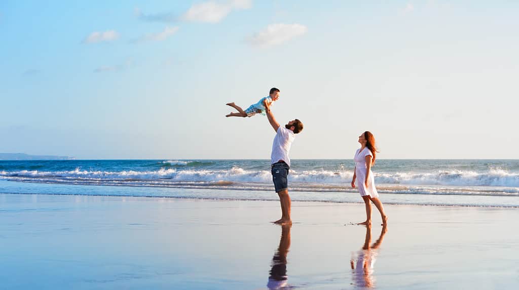 Happy family holidays. Joyful father, mother, baby son walk with fun along edge of sunset sea surf on black sand beach. Active parents and people outdoor activity on summer vacations with children.