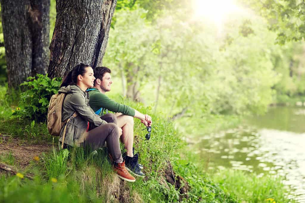 travel, hiking, backpacking, tourism and people concept - smiling couple with backpacks resting on river bank in nature on river bank