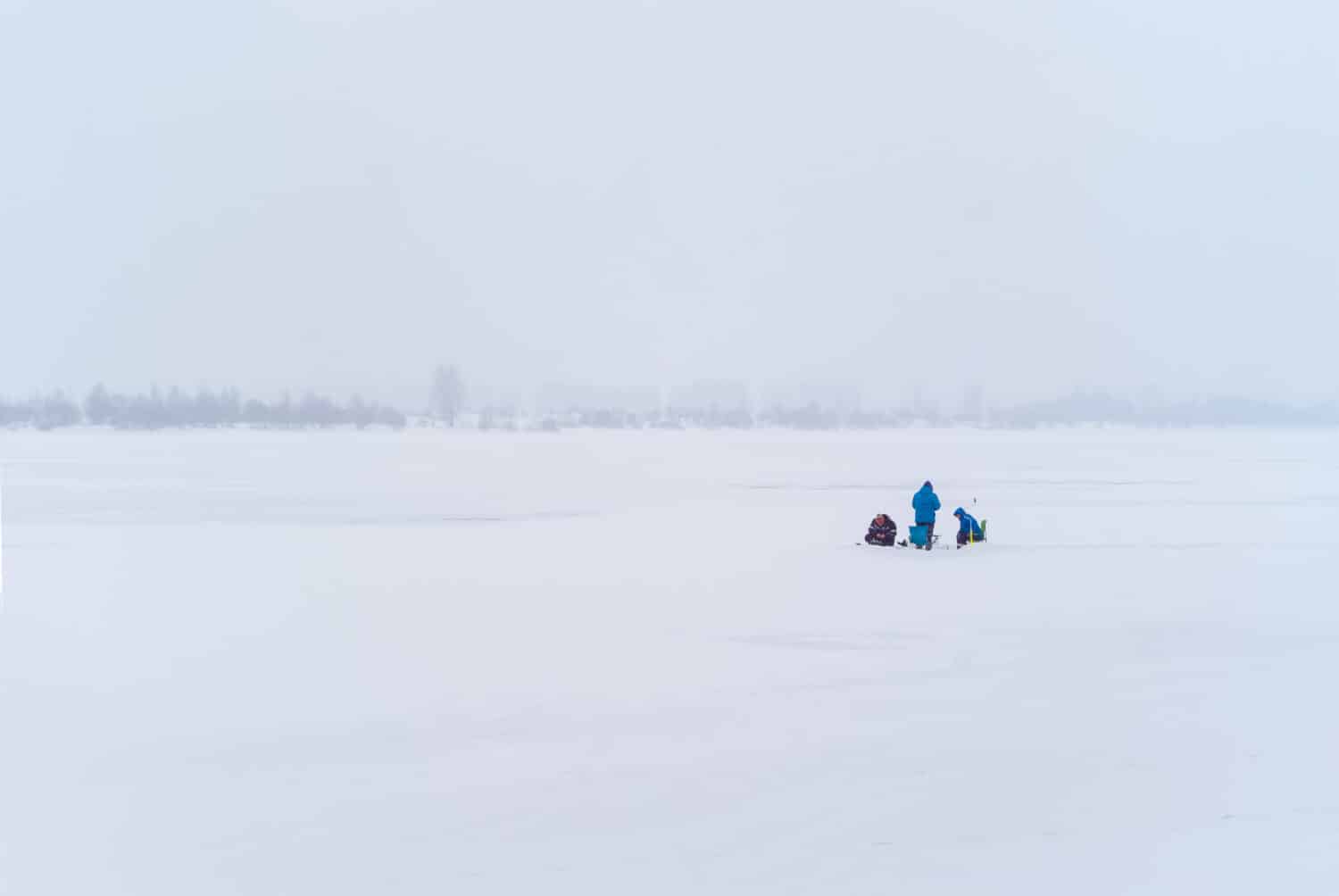 three fishermen in the distance do ice fishing in a foggy snowy winter landscape