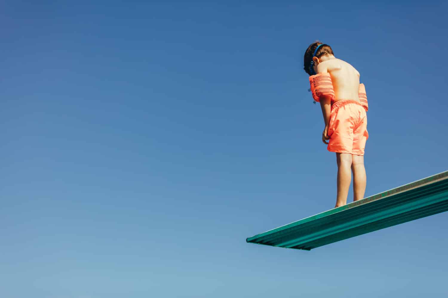 Low angle shot of boy with sleeves floats on diving board preparing for dive in the pool. Boy standing on diving spring board against sky.