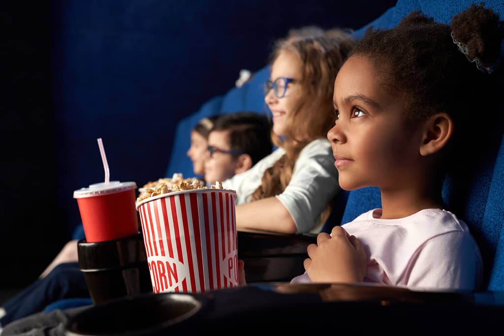 Beautiful african girl with funny hairstyle watching excited movie in cinema. Wonderful little child sitting with friends, eating popcorn and smiling. Concept of entertainment and enjoyment.