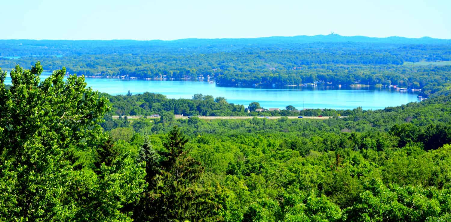 A view of Pewaukee Lake surrounded by deep green woodlands from high above with a highway running alongside the distant waters. 